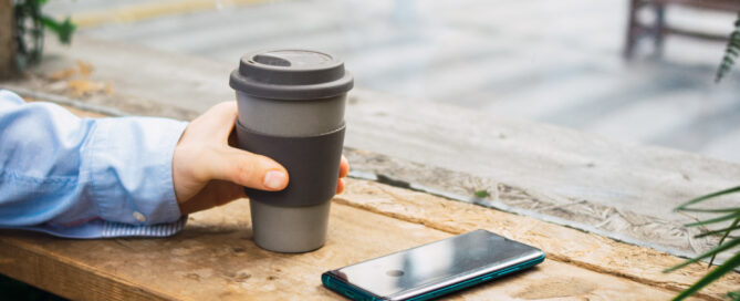 Person holding re-usable coffee cup and phone on the table.